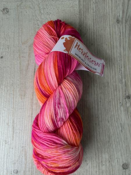 Handgefärbter Wollstrang „Neonherz“ von Herzfasern auf der Qualität No. 21. Leuchtende Neonpink-, Korall- und Orangetöne verlaufen weich ineinander. Das Garn liegt auf hellem Holzhintergrund und zeigt intensive, moderne Farbkontraste.